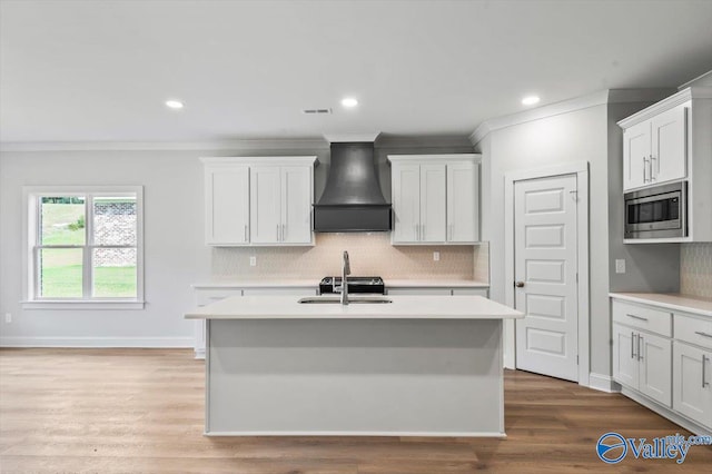 kitchen featuring a kitchen island with sink, hardwood / wood-style floors, premium range hood, stainless steel microwave, and white cabinets