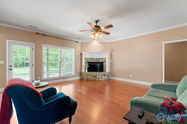 living room with a ceiling fan, wood finished floors, crown molding, a stone fireplace, and baseboards