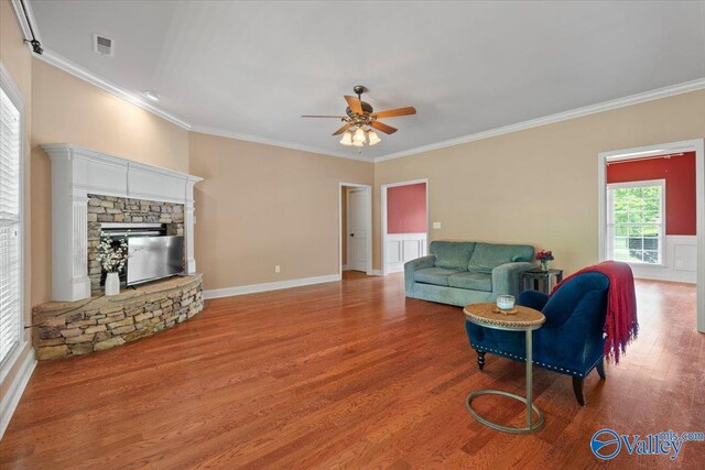 living room featuring ceiling fan, wood finished floors, crown molding, baseboards, and a stone fireplace