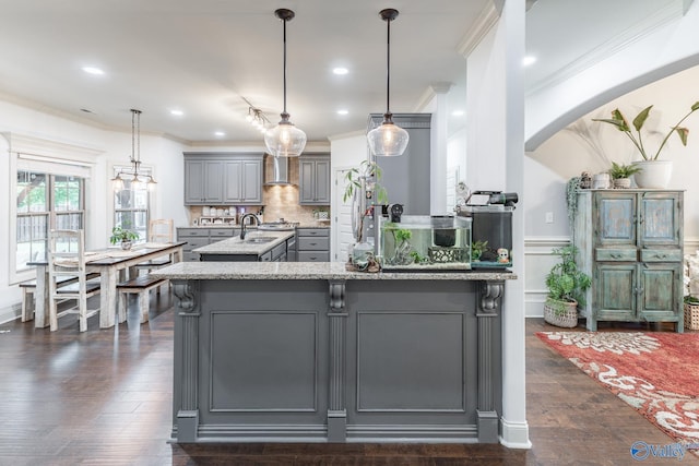 kitchen with ornamental molding, an island with sink, gray cabinets, light stone counters, and arched walkways