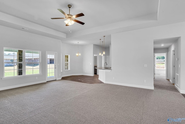 unfurnished living room with a tray ceiling, ceiling fan with notable chandelier, baseboards, and dark carpet