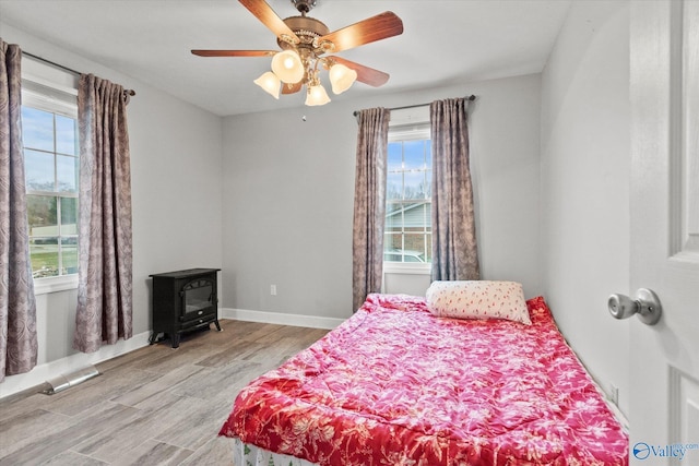 bedroom with ceiling fan, a wood stove, multiple windows, and light hardwood / wood-style flooring