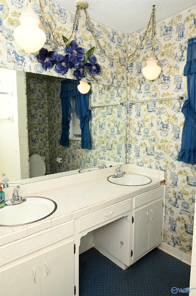 full bath with double vanity, wallpapered walls, a textured ceiling, and a sink