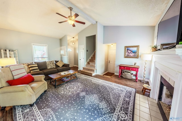 tiled living room featuring a ceiling fan, a fireplace, baseboards, vaulted ceiling with beams, and stairs