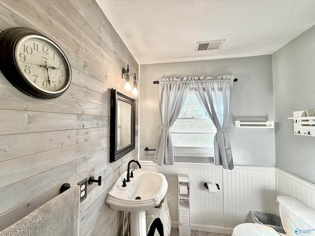 half bathroom with a textured ceiling, toilet, a wainscoted wall, a sink, and visible vents