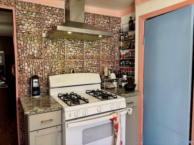 kitchen with dark stone countertops, gas range gas stove, crown molding, and wall chimney range hood