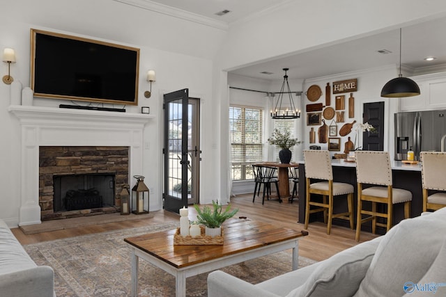 living area with light wood-type flooring, visible vents, ornamental molding, a stone fireplace, and an inviting chandelier