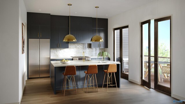 kitchen featuring light hardwood / wood-style flooring, tasteful backsplash, hanging light fixtures, stainless steel built in refrigerator, and an island with sink