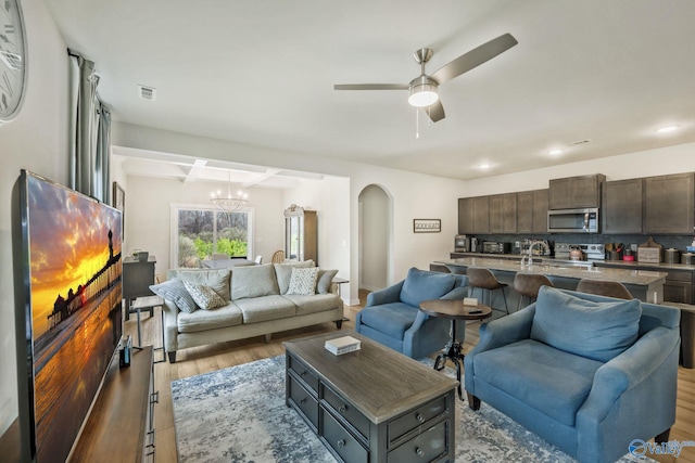 living area featuring visible vents, arched walkways, beamed ceiling, light wood-type flooring, and ceiling fan with notable chandelier