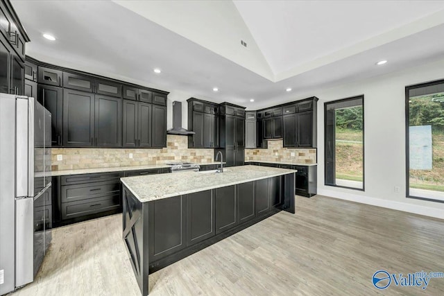 kitchen with light stone counters, stainless steel appliances, backsplash, wall chimney exhaust hood, and an island with sink