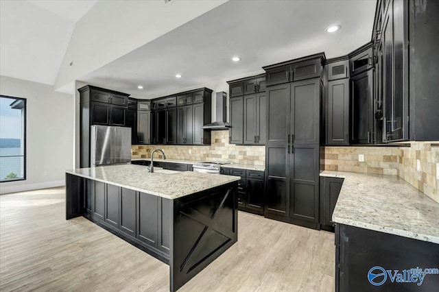 kitchen with a center island with sink, wall chimney exhaust hood, appliances with stainless steel finishes, light stone counters, and dark cabinetry