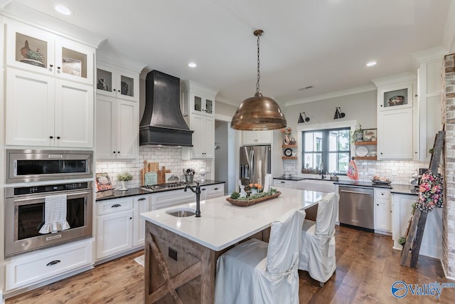 kitchen featuring custom exhaust hood, wood-type flooring, an island with sink, appliances with stainless steel finishes, and backsplash