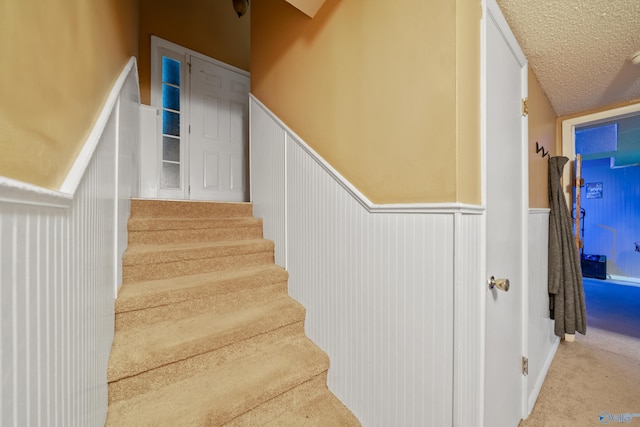 stairway with a textured ceiling, carpet floors, and wainscoting