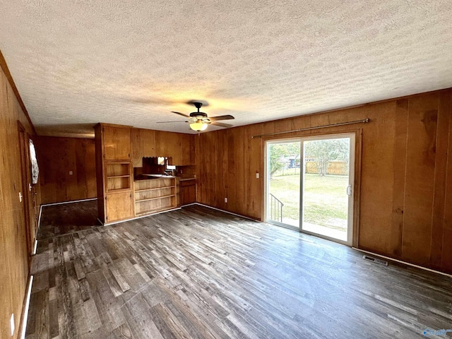 unfurnished living room with visible vents, wooden walls, ceiling fan, dark wood finished floors, and a textured ceiling