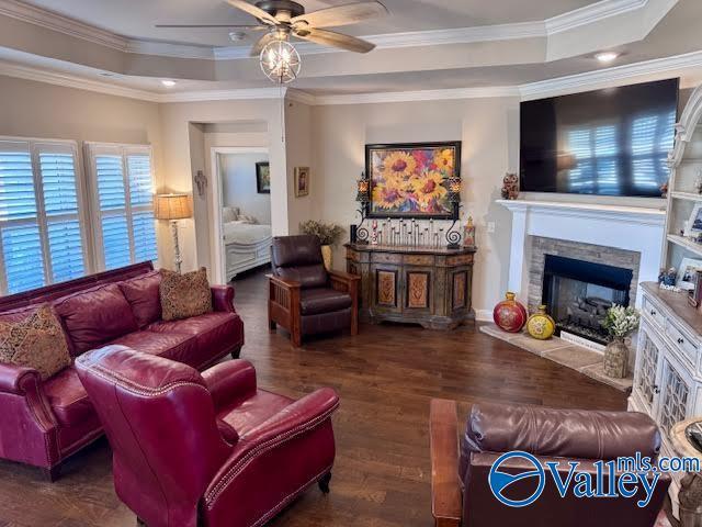 living room featuring a stone fireplace, ornamental molding, a tray ceiling, and wood finished floors