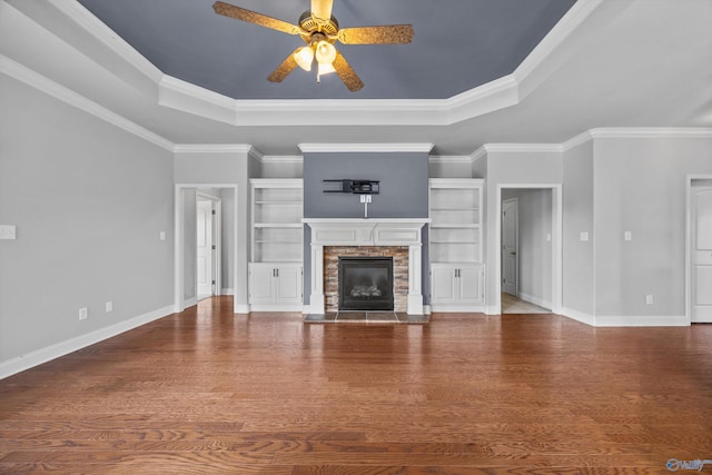 unfurnished living room with ceiling fan, a stone fireplace, wood finished floors, baseboards, and a tray ceiling