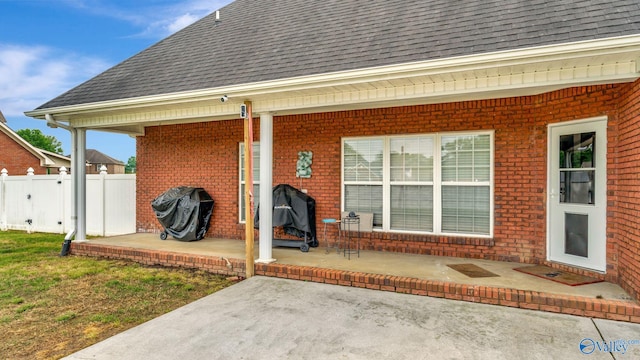 view of exterior entry featuring fence, a shingled roof, a patio area, and brick siding