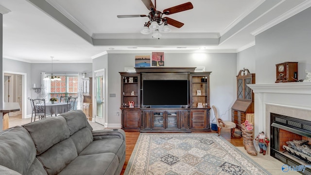 living area with a fireplace with flush hearth, a raised ceiling, light wood-style flooring, ornamental molding, and ceiling fan with notable chandelier