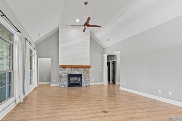 unfurnished living room with high vaulted ceiling, ceiling fan, light wood-type flooring, a stone fireplace, and baseboards