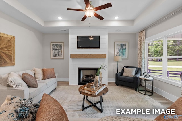 living room with ceiling fan, light wood-type flooring, a fireplace, and a raised ceiling