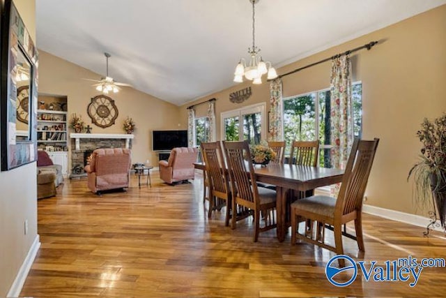 dining space featuring baseboards, vaulted ceiling, light wood-type flooring, a fireplace, and ceiling fan with notable chandelier