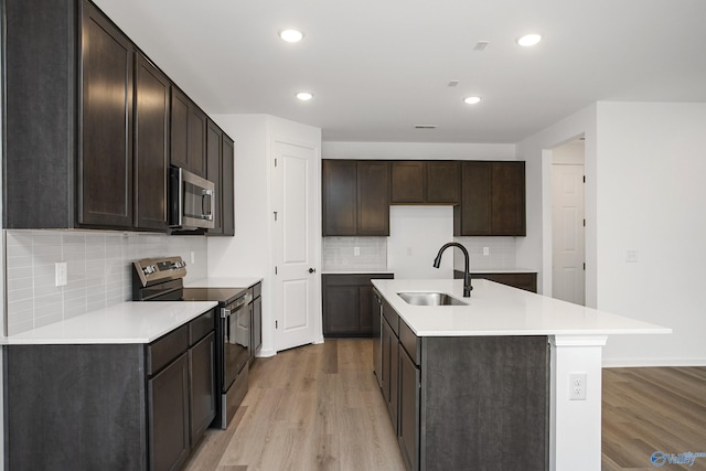 kitchen with sink, a kitchen island with sink, stainless steel appliances, light hardwood / wood-style floors, and decorative backsplash