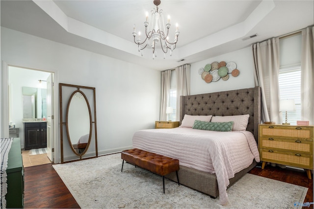 bedroom with a raised ceiling, visible vents, ensuite bathroom, wood finished floors, and a chandelier