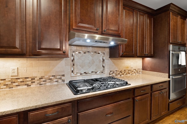 kitchen featuring black gas stovetop, stainless steel double oven, light stone counters, under cabinet range hood, and light wood-style floors