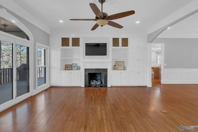 unfurnished living room featuring built in features, wood finished floors, a fireplace with flush hearth, arched walkways, and crown molding