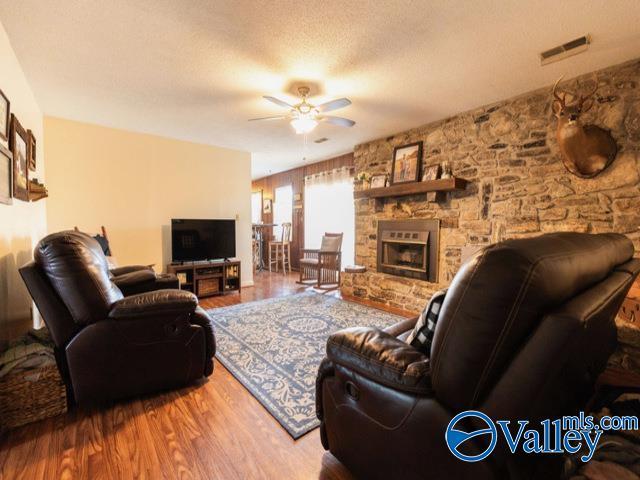 living room featuring wood-type flooring, a textured ceiling, a stone fireplace, and ceiling fan