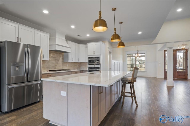 kitchen with premium range hood, white cabinetry, hanging light fixtures, appliances with stainless steel finishes, and a center island with sink