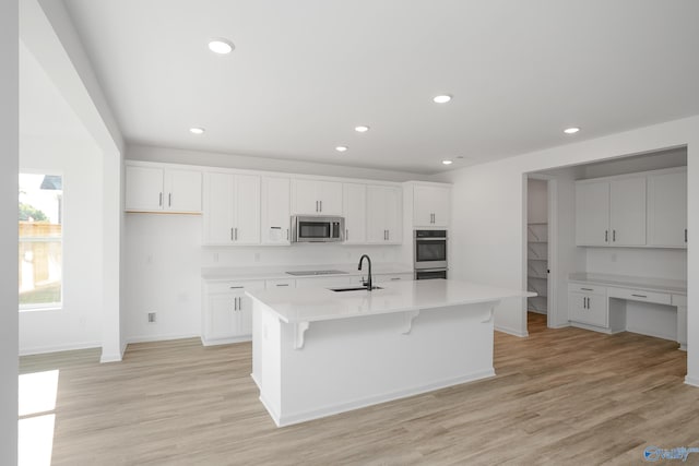 kitchen featuring built in desk, an island with sink, sink, white cabinets, and light wood-type flooring