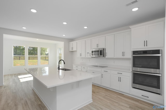 kitchen with appliances with stainless steel finishes, an island with sink, sink, white cabinets, and light hardwood / wood-style floors