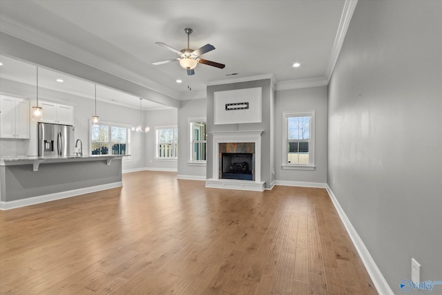 unfurnished living room featuring a tile fireplace, ceiling fan with notable chandelier, baseboards, light wood-type flooring, and crown molding