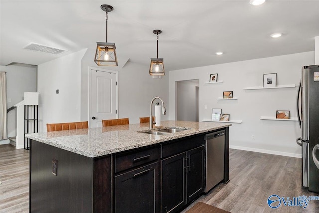 kitchen with appliances with stainless steel finishes, a kitchen island with sink, light stone countertops, pendant lighting, and sink