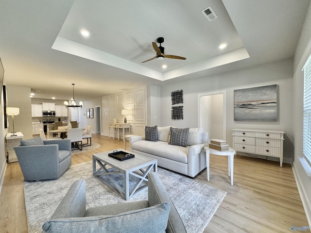 living room with light wood-type flooring, a tray ceiling, and ceiling fan with notable chandelier