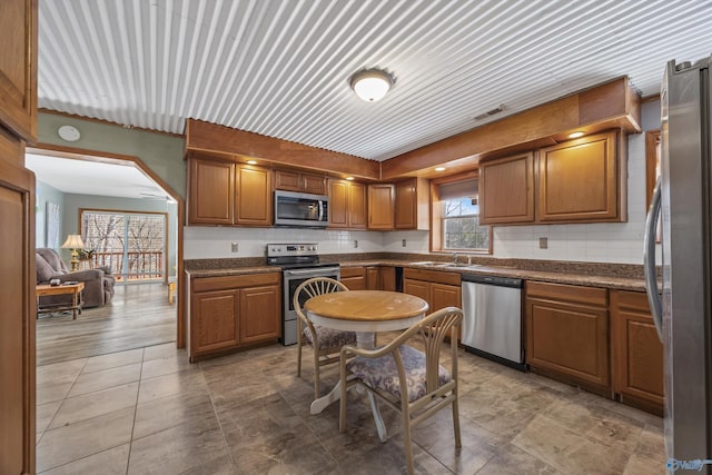 kitchen featuring stainless steel appliances, dark countertops, backsplash, brown cabinetry, and a sink