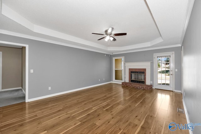 unfurnished living room featuring ornamental molding, wood-type flooring, a tray ceiling, ceiling fan, and a brick fireplace