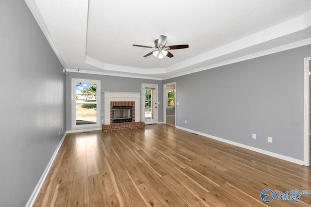 unfurnished living room with ceiling fan, a tray ceiling, wood-type flooring, a fireplace, and crown molding