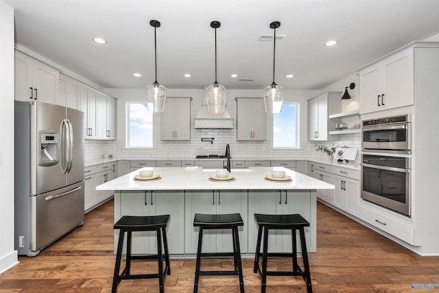 kitchen featuring appliances with stainless steel finishes, dark hardwood / wood-style flooring, custom range hood, pendant lighting, and an island with sink