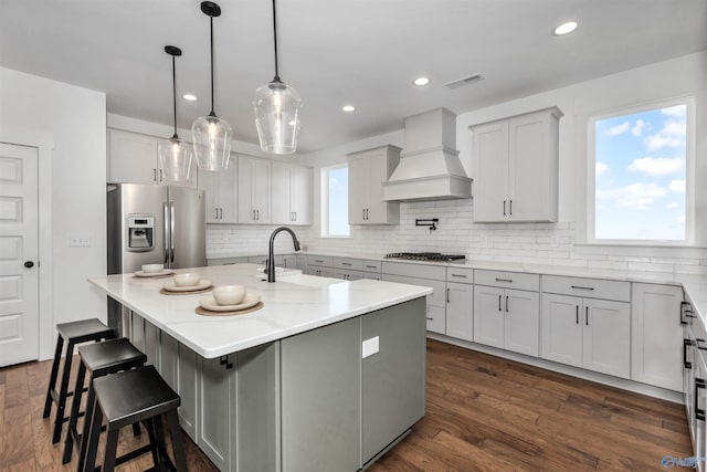 kitchen featuring appliances with stainless steel finishes, premium range hood, sink, pendant lighting, and a center island with sink