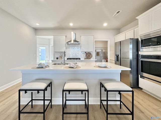 kitchen featuring wall chimney exhaust hood, light wood-type flooring, a center island with sink, and appliances with stainless steel finishes