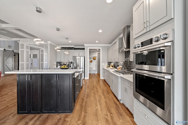 kitchen featuring a kitchen island with sink, wall chimney range hood, stainless steel appliances, decorative light fixtures, and decorative backsplash