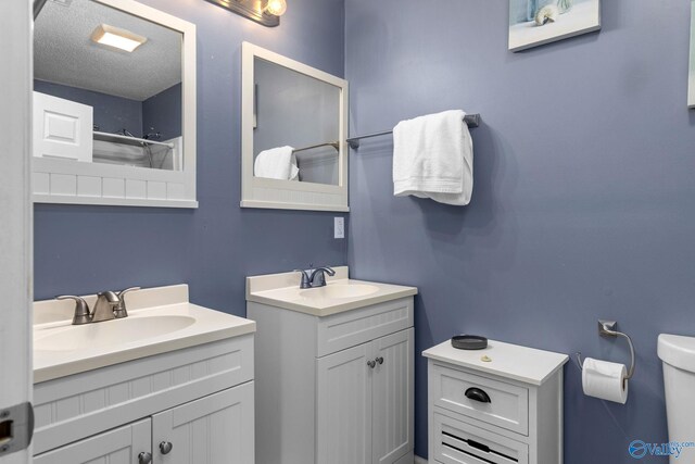 full bath featuring toilet, a textured ceiling, two vanities, and a sink