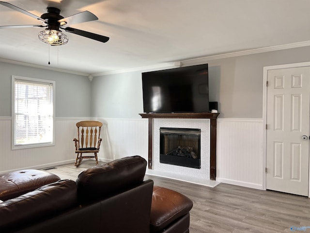 living room featuring a tiled fireplace, wainscoting, ceiling fan, ornamental molding, and wood finished floors