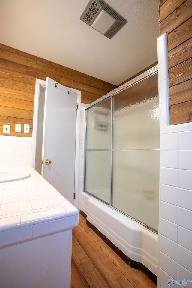 full bath featuring combined bath / shower with glass door, wood walls, wood finished floors, and visible vents