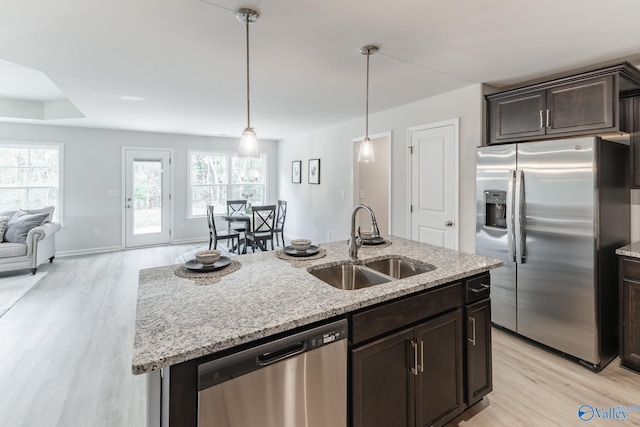 kitchen with sink, appliances with stainless steel finishes, dark brown cabinets, light stone counters, and an island with sink