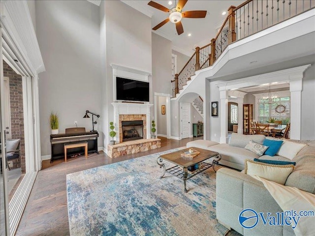 living room featuring a stone fireplace, wood finished floors, baseboards, ceiling fan with notable chandelier, and a high ceiling
