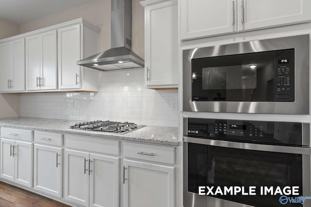 kitchen with wall chimney exhaust hood, white cabinetry, appliances with stainless steel finishes, light stone counters, and light hardwood / wood-style floors