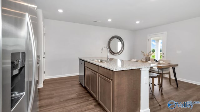 kitchen featuring appliances with stainless steel finishes, sink, light stone countertops, dark wood-type flooring, and a center island with sink